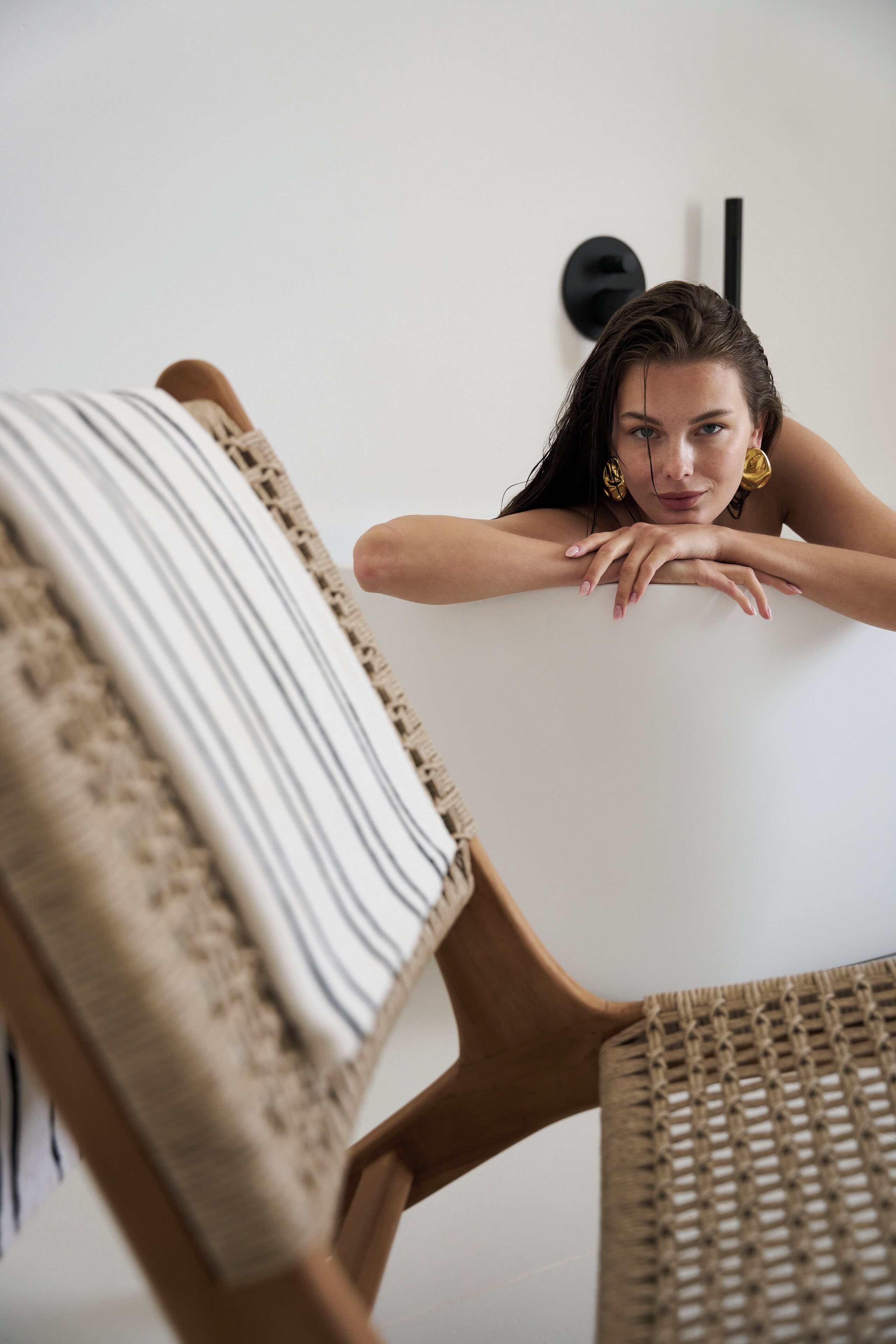 A person leaning on a white wall with a woven chair and a wooden chair with a striped cushion.