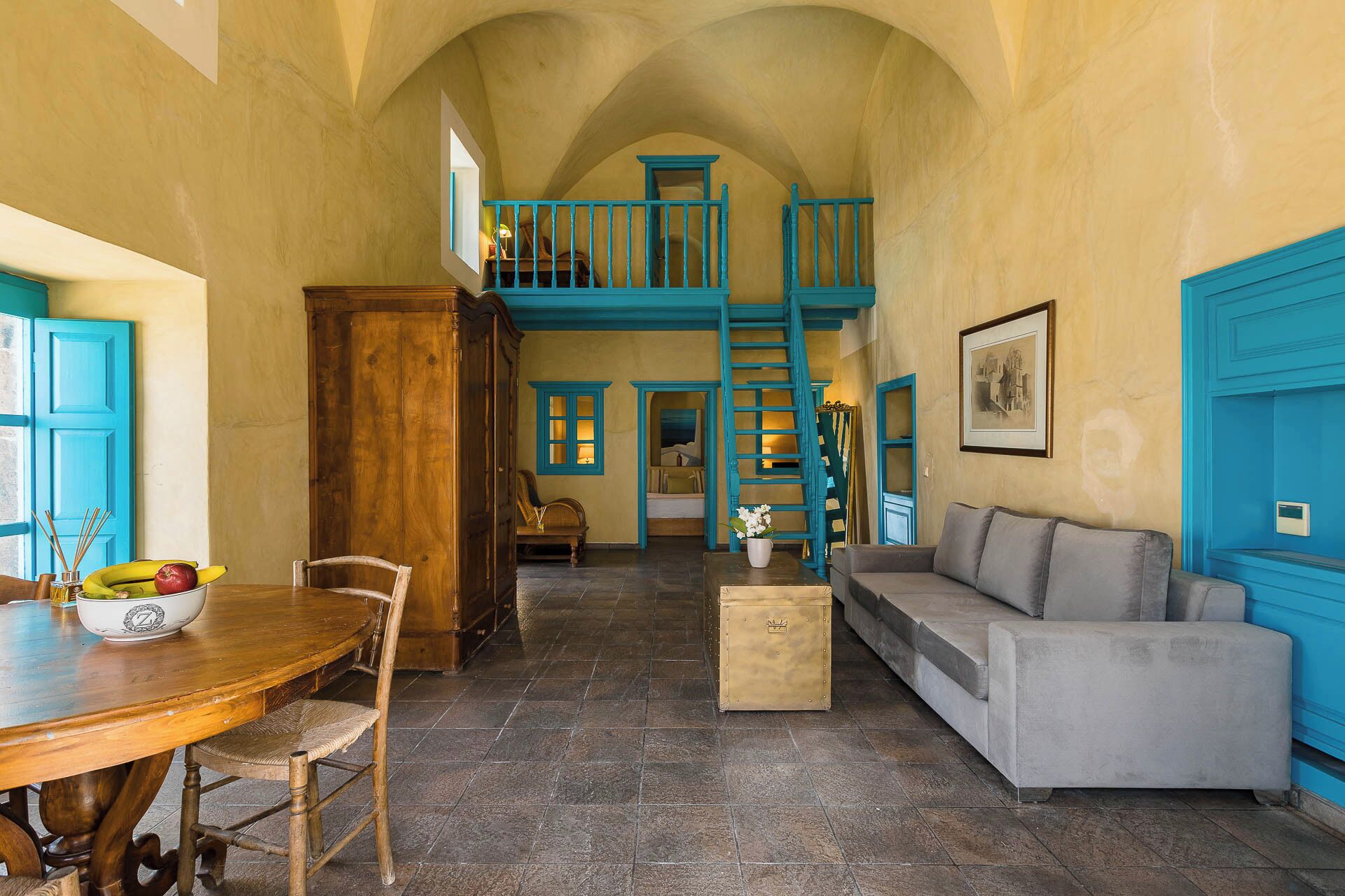 A spacious room with a high ceiling, a staircase, a dining area with a wooden table and chairs, a sofa, and a small table with a vase.