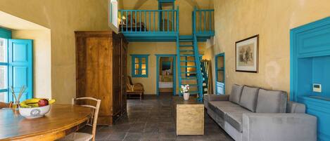 A spacious room with a high ceiling, a staircase, a dining area with a wooden table and chairs, a sofa, and a small table with a vase.