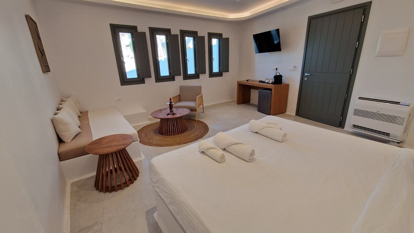 A modern hotel room with a bed, a sofa, a small round table, a desk, a TV, and a window with shutters.