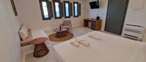A modern hotel room with a bed, a sofa, a small round table, a desk, a TV, and a window with shutters.