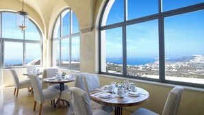 A dining area with tables set for four, large arched windows, and a view of a coastal landscape. - Zannos Melathron Hotel by Omilos Hotels (Santorini)