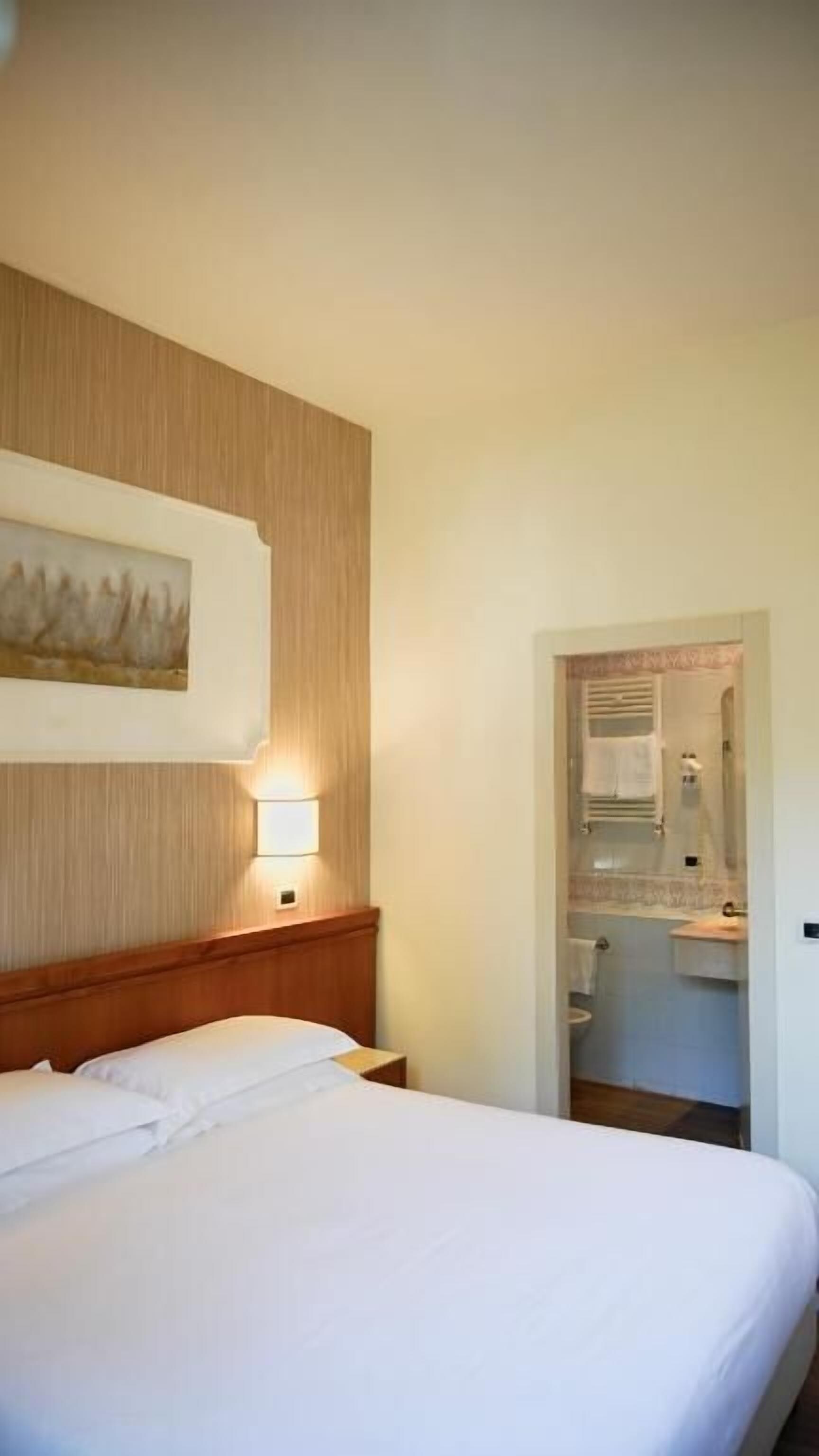A hotel room with a bed, a nightstand, a wall-mounted lamp, and a painting. There is a bathroom visible through an open door.