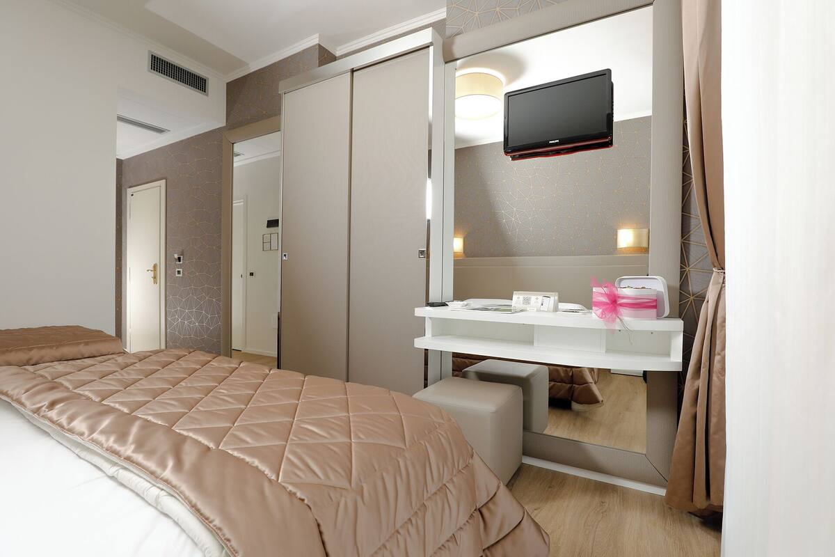 A hotel room with a bed, a vanity with a mirror, a flat-screen TV, and a bedside table with a lamp and a gift box.