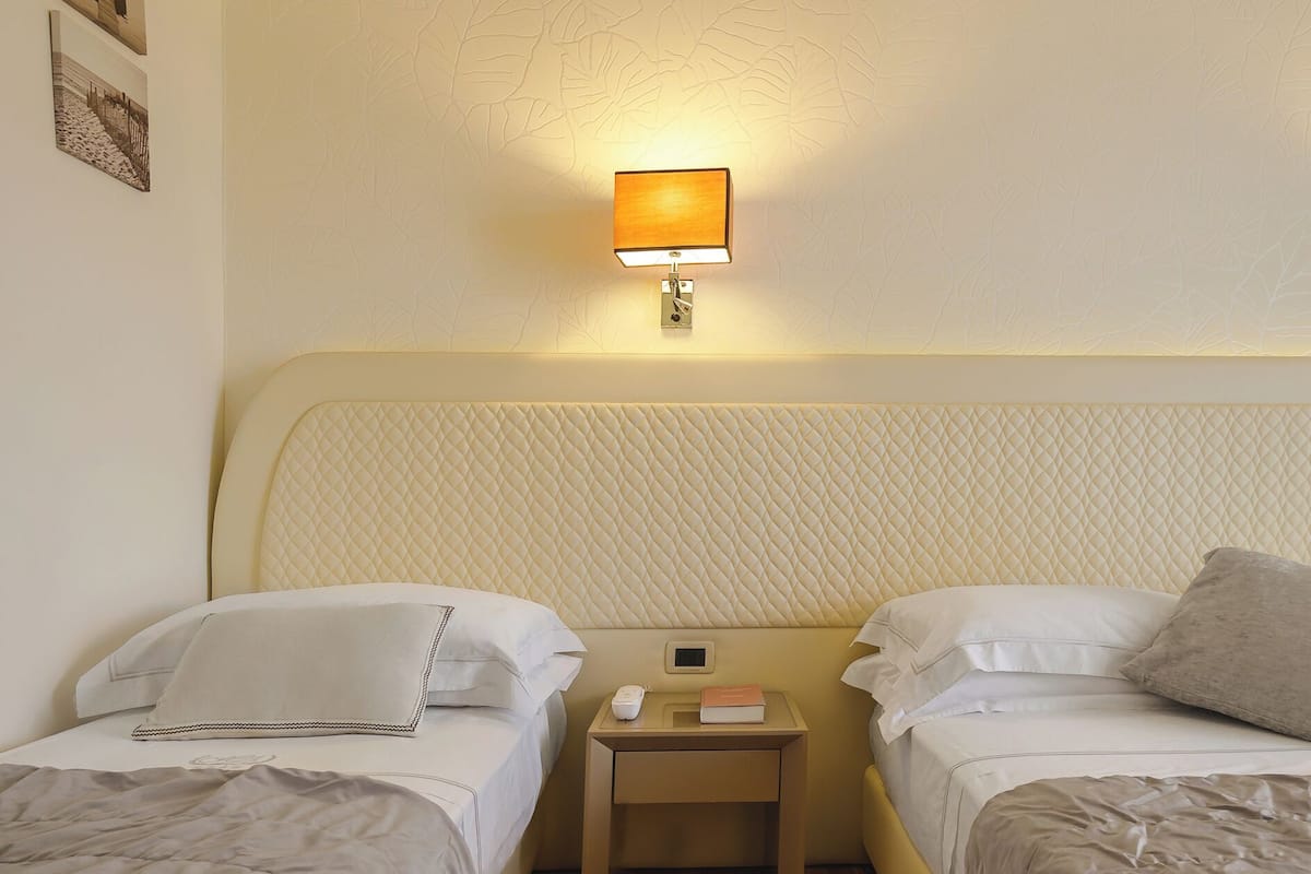 A hotel room with two beds, a bedside table with a lamp, a phone, and a book.