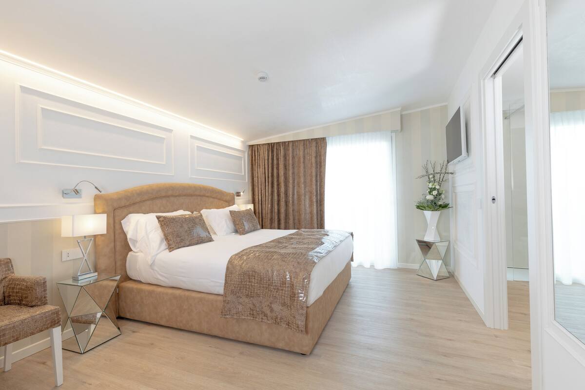 A modern bedroom with a large bed, a bedside table with a lamp, and a view of the outdoors through a window.