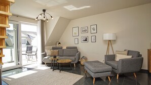 A modern living room with a grey sofa, a small coffee table, and a lamp. There are framed pictures on the wall and a staircase leading to an upper floor. - Geräumige Ferienwohnung Absoluter Strandnähe - Haus Sanddorn Fewo 07 (Thiessow)