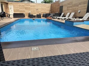 Una piscina rectangular con borde de azulejos, rodeada de sillas de descanso y una terraza de madera.