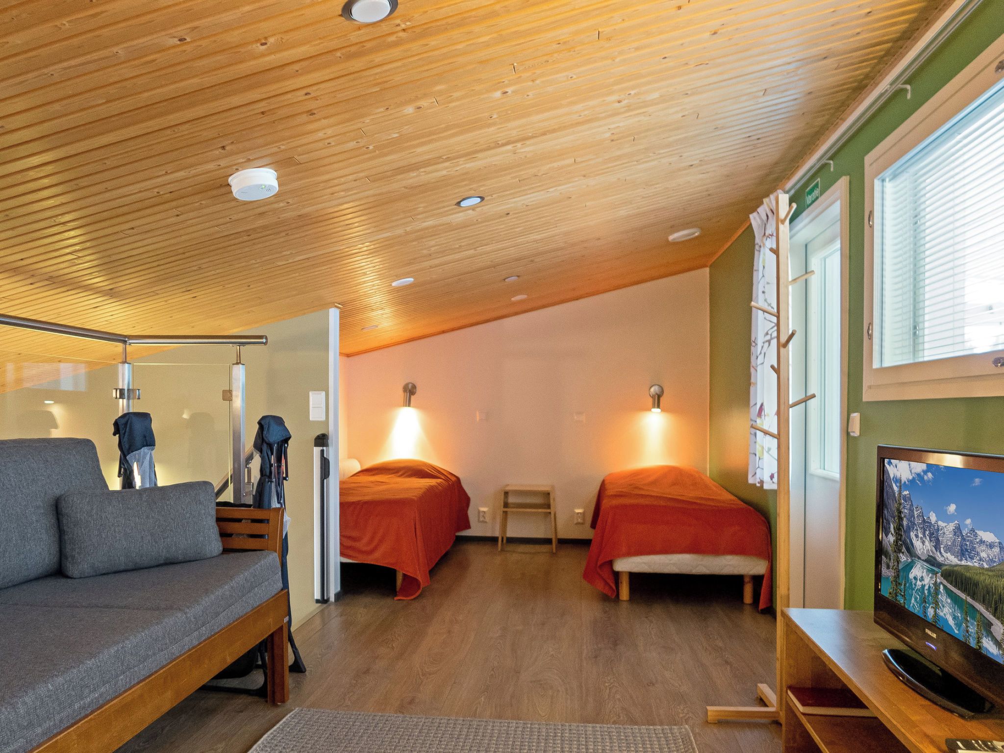 A compact room with a wooden ceiling, two beds with red bedding, a grey sofa, a small table, and a television on a wooden stand.