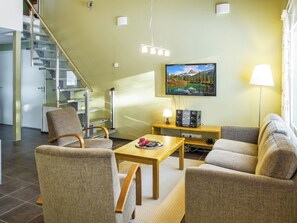 A modern living room with a sofa, armchairs, a coffee table, and a television. There is a staircase leading to an upper level.