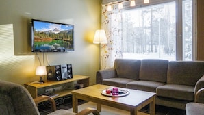 A cozy living room with a flat-screen TV, a sofa, a coffee table with a tray of items, and a window with blinds.