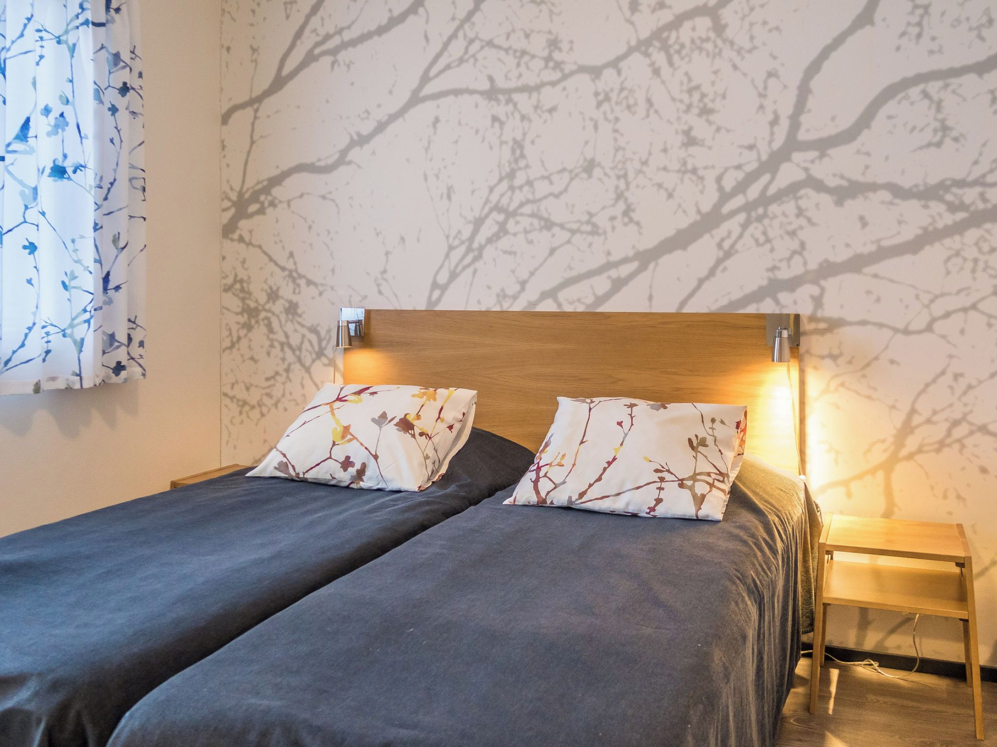 A double bed with two matching pillows, a wooden headboard, a bedside table with a lamp, and a wall with a tree branch pattern.