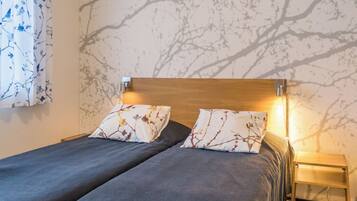 A double bed with two matching pillows, a wooden headboard, a bedside table with a lamp, and a wall with a tree branch pattern.