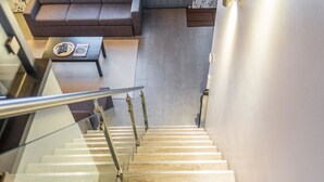 Escadaria moderna com degraus de madeira e guarda-corpos de vidro, que dá acesso a uma sala de estar com sofá e mesa de centro.