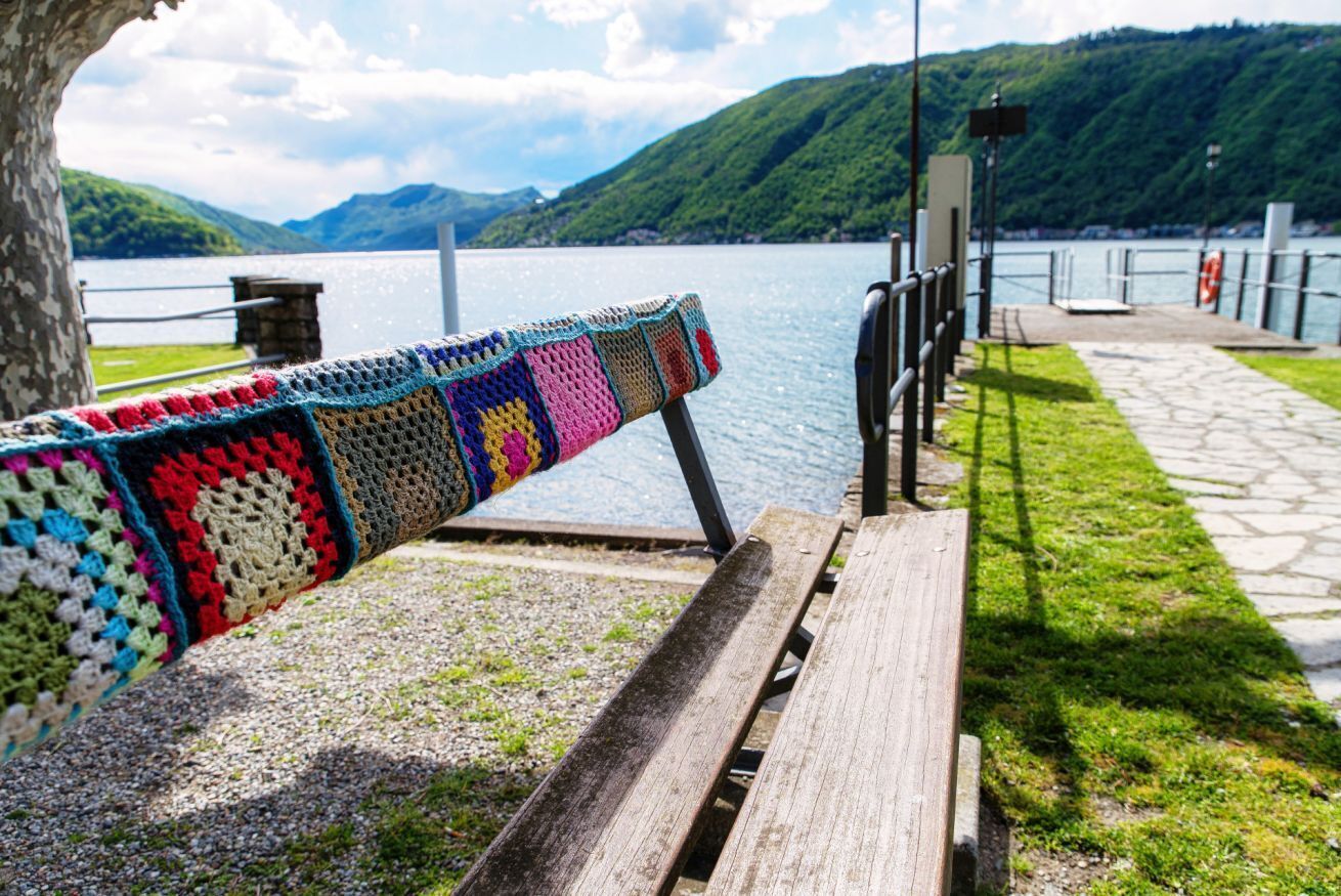 A wooden bench with a colorful crocheted blanket draped over it, overlooking a lake with mountains in the background.