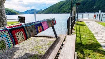 A wooden bench with a colorful crocheted blanket draped over it, overlooking a lake with mountains in the background.