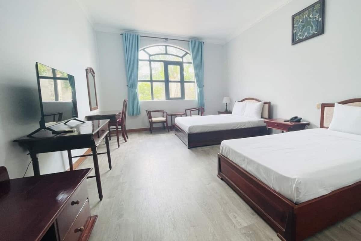 Une chambre d’hôtel avec deux lits simples, un bureau avec une chaise, une petite table et une fenêtre avec des rideaux.