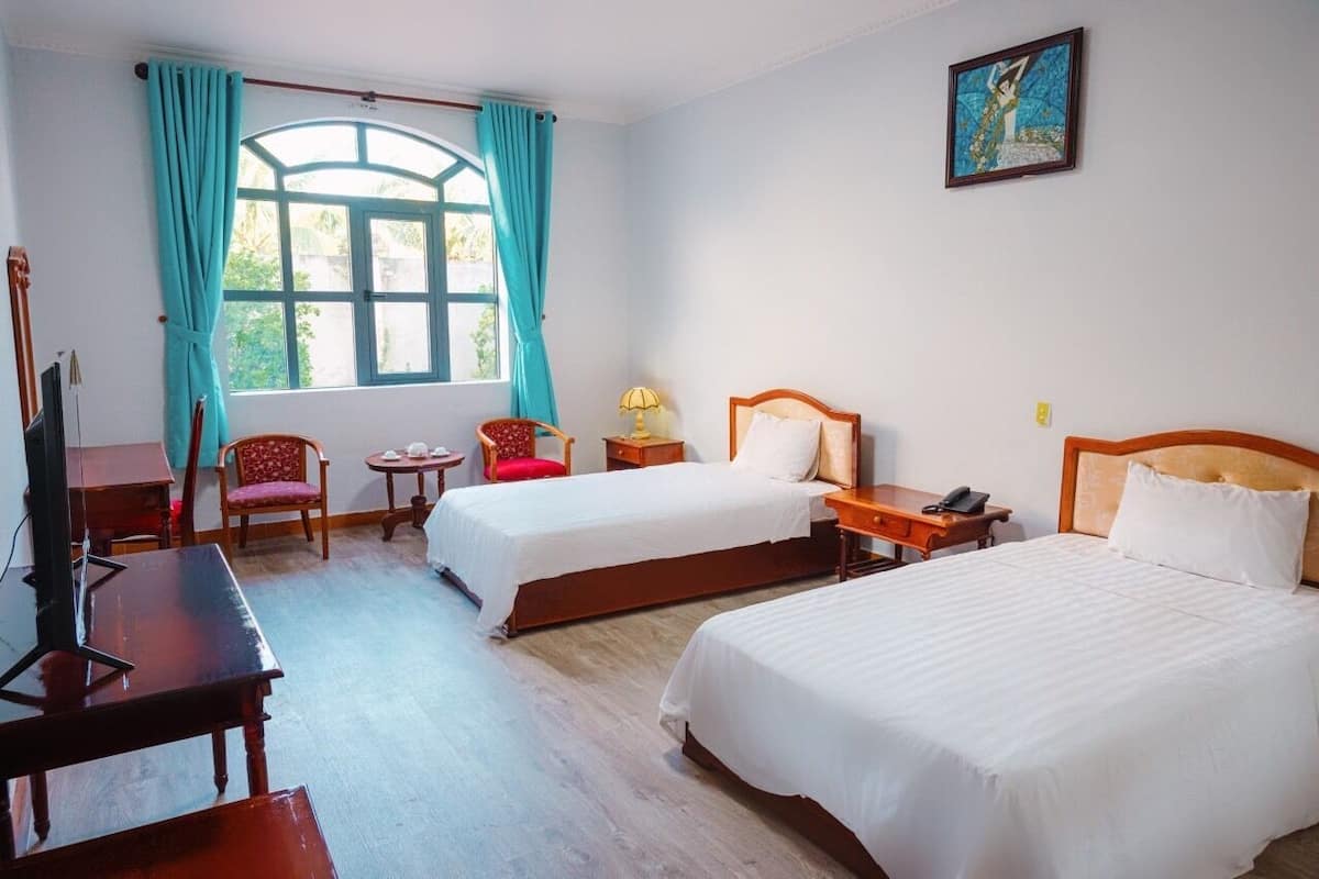 Une chambre d’hôtel avec deux lits, un bureau, une chaise, une petite table et une fenêtre avec des rideaux.