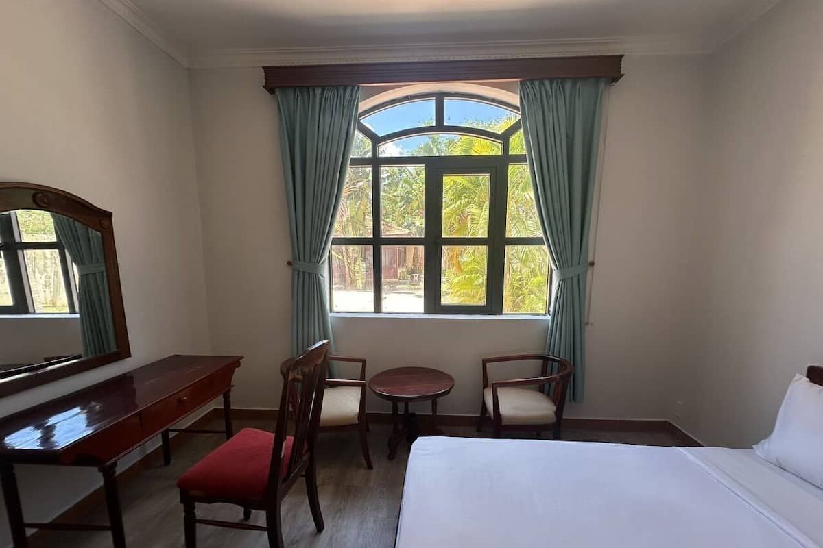 Une chambre d’hôtel comprenant un lit, un bureau, deux chaises, une table ronde et une grande fenêtre avec des rideaux.