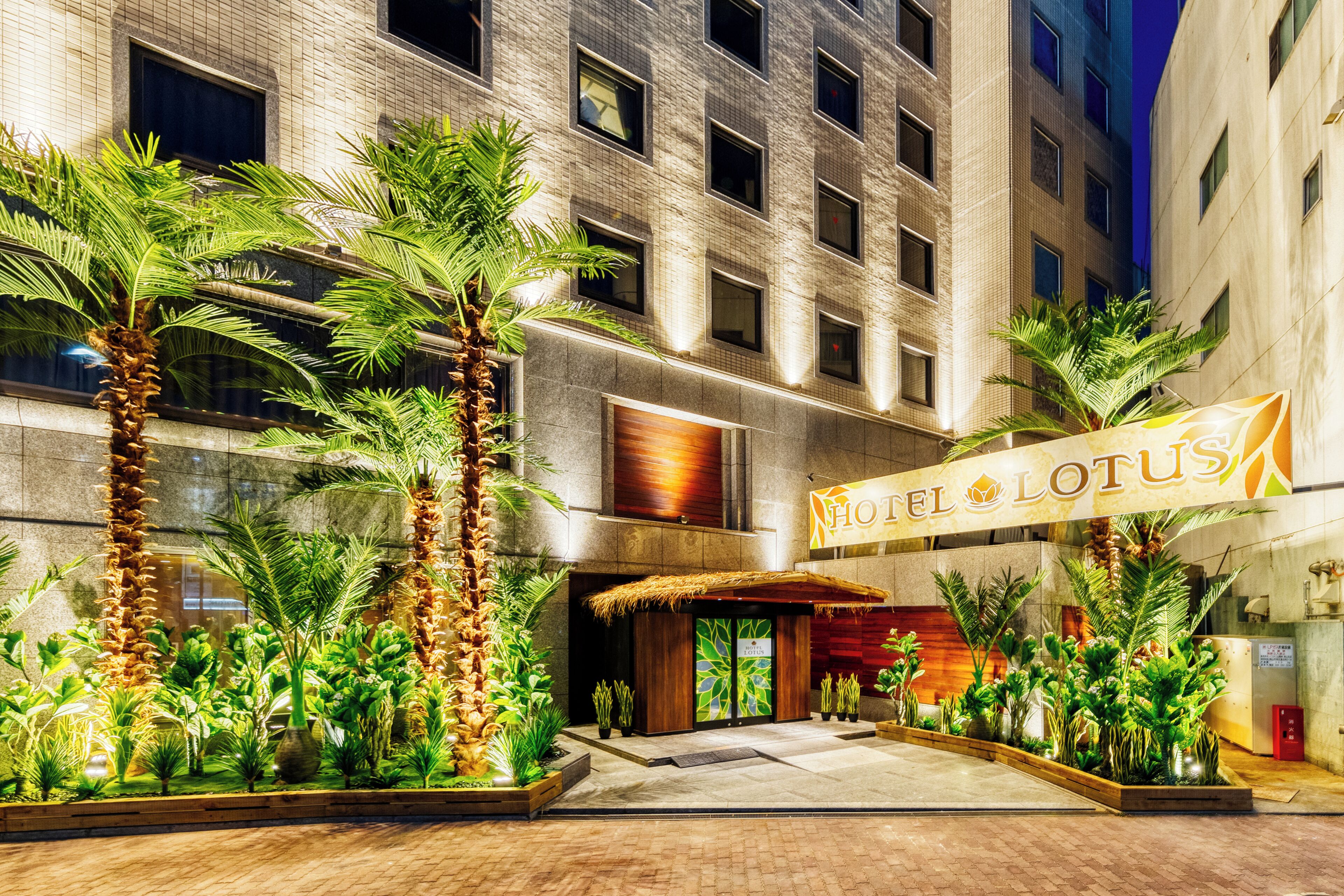 Entrada de un hotel con un letrero que dice 'HOTEL LOTUS', rodeada de palmeras y fachada de un edificio de ladrillo.