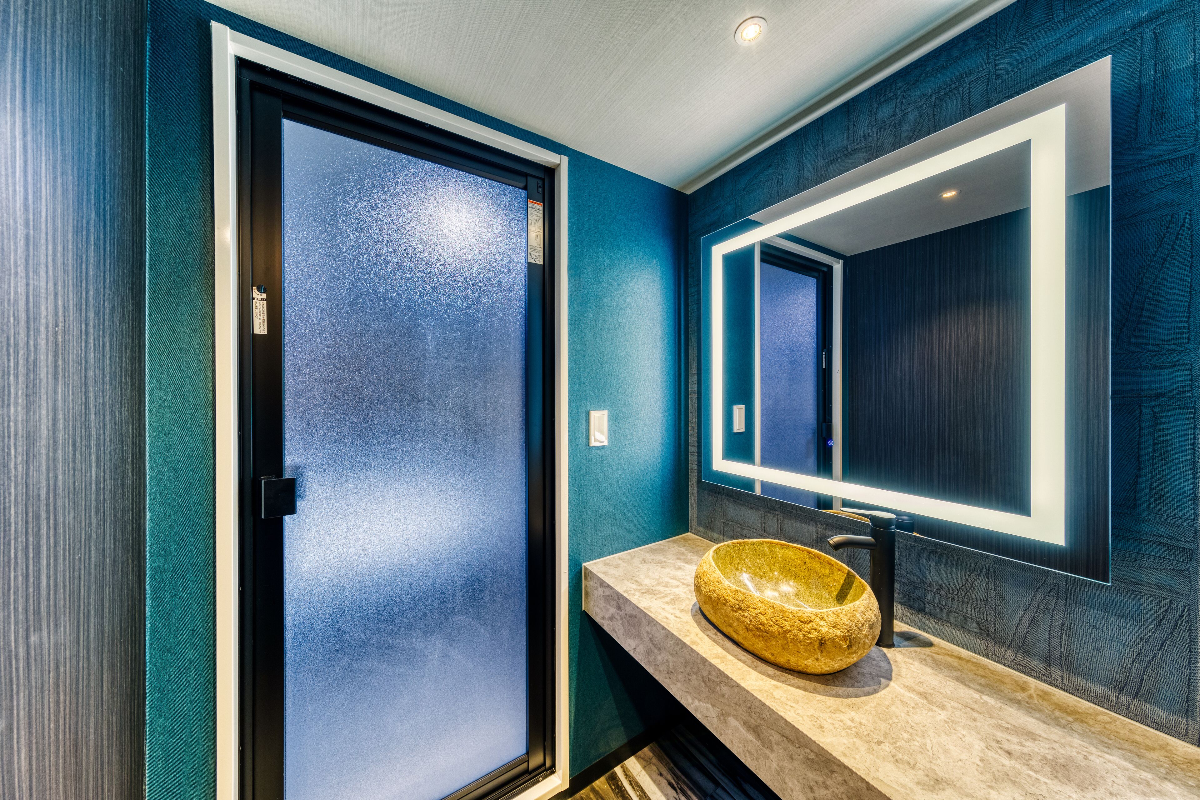 Une salle de bain moderne avec un grand miroir, un lavabo en pierre et une porte en verre dépoli.