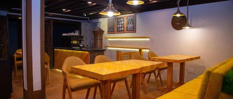 Interior de un restaurante con mesas y sillas de madera, lámparas colgantes y una barra de fondo.