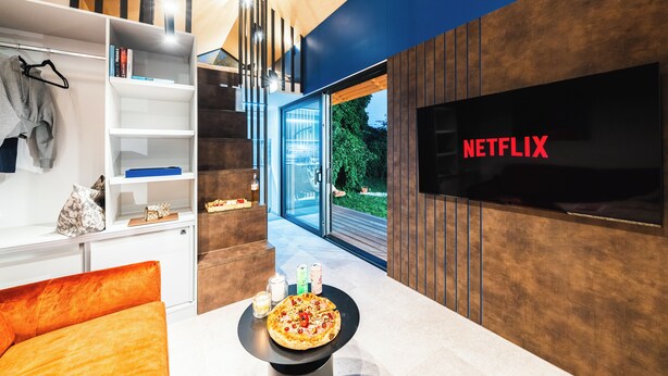 A modern living room with a pizza on a table, a bookshelf, a TV displaying "NETFLIX", and a staircase leading to a glass door. - Ostravinka - Boutique Apartment (Ostrava)