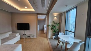 Una habitación de hotel moderna con televisión de pantalla plana, un sofá, una mesa de comedor con sillas, una planta en maceta y un amplio ventanal con vistas a la ciudad.