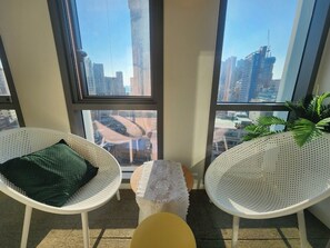 Een moderne kamer met grote ramen, twee witte rieten stoelen, een klein tafeltje en uitzicht op de skyline.
