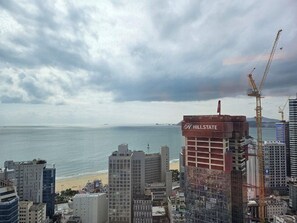 Een kuststadsgezicht met een opvallend HillState gebouw in aanbouw, omringd door hoge flats en een strand.
