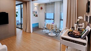 Una habitación de hotel moderna con suelo de madera, una zona de comedor con mesa y sillas, un televisor montado en la pared y una ventana con persianas.