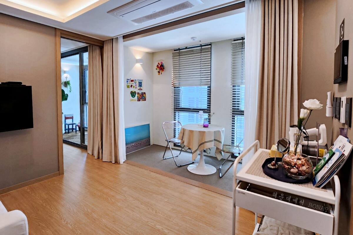 Ein modernes Hotelzimmer mit Holzboden, einer Essecke mit Tisch und Stühlen, einem an der Wand montierten Fernseher und einem Fenster mit Jalousien.