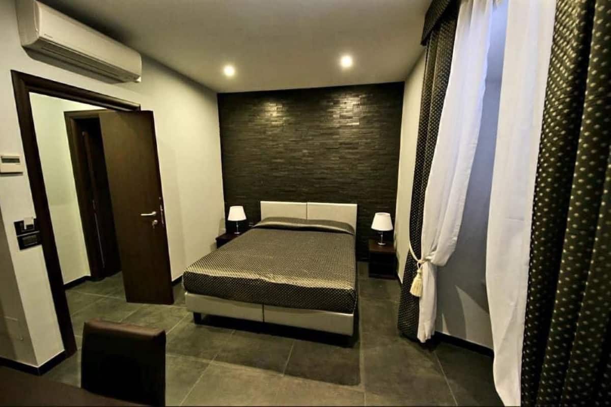 A modern hotel room with a large bed, two bedside tables with lamps, a dark textured wall, and a door leading to another room.