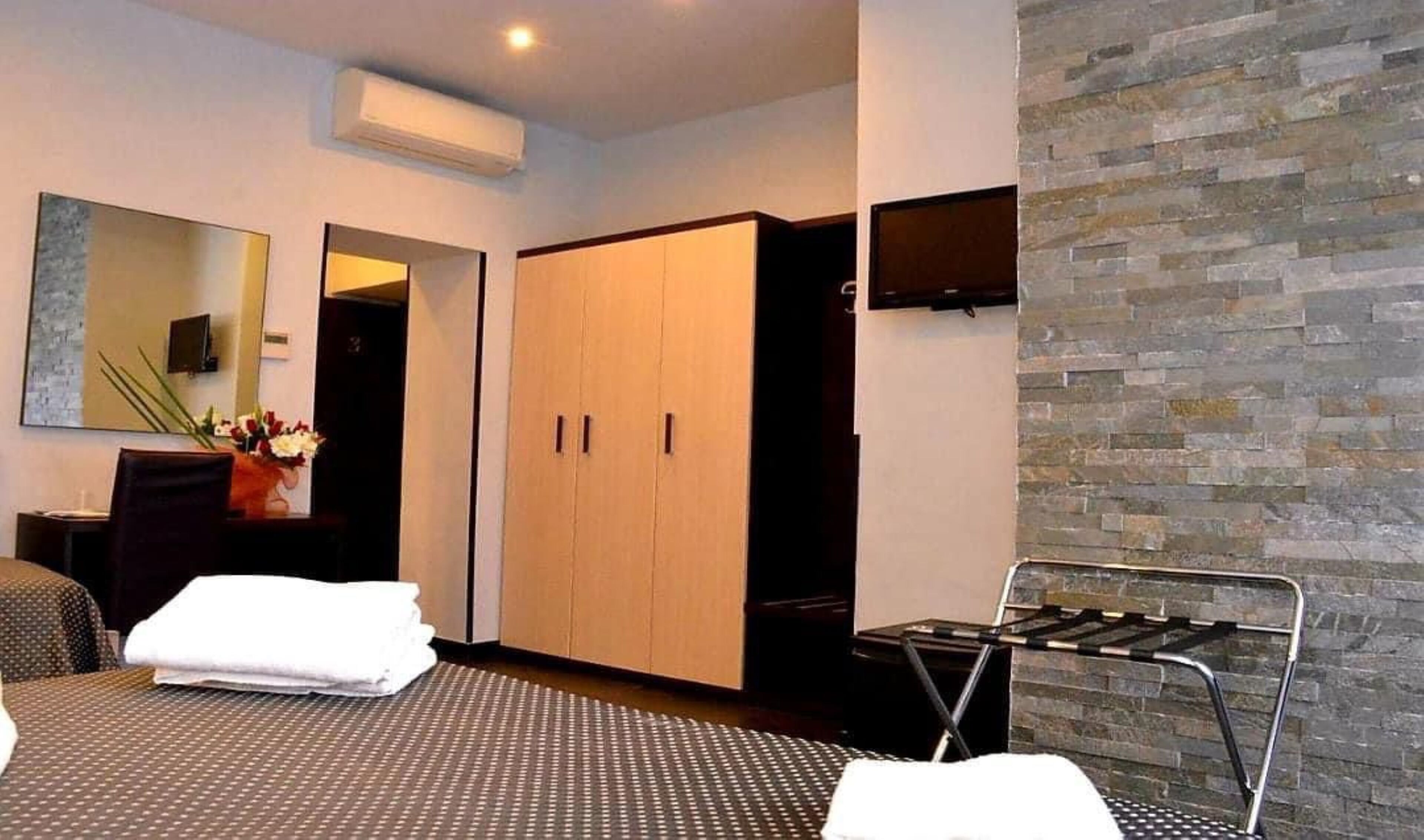 A modern hotel room with a bed, a seating area with a table and chairs, a wardrobe, and a stone wall.