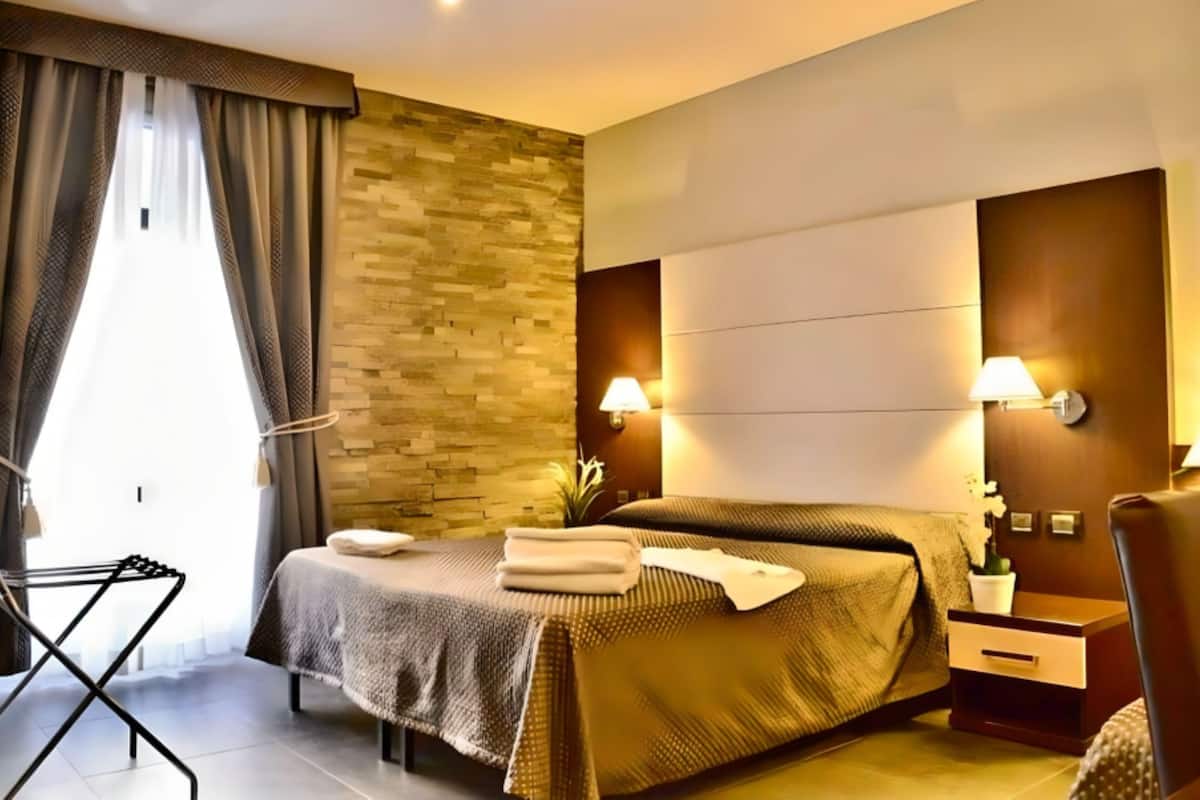 A modern hotel room with a bed, bedside tables, a wall-mounted lamp, and a window with curtains.