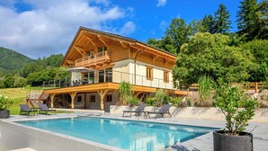 A wooden chalet with a pool, surrounded by greenery and hills.