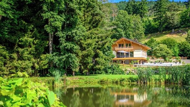 A wooden house with a balcony, surrounded by dense forest and a pond.