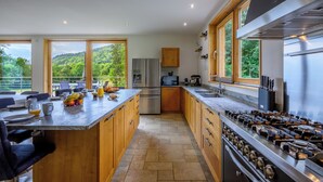 A modern kitchen with a large island, stainless steel appliances, wooden cabinets, and large windows offering a view of the outdoors.