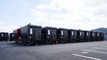 Une rangée de petits bâtiments modulaires noirs, dotés de portes numérotées, est disposée sur un parking, sous un ciel dégagé et avec des collines qui se dessinent à l’horizon.