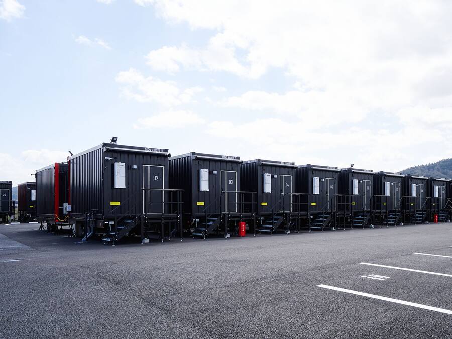 Une rangée de petits bâtiments modulaires noirs, dotés de portes numérotées, est disposée sur un parking, sous un ciel dégagé et avec des collines qui se dessinent à l’horizon.