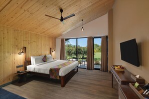 A hotel room with a wooden ceiling, a bed with a colorful blanket, a desk with a flat-screen TV, and a balcony with a view of greenery. - Soil to Soul Resort - Statue of Unity (Tilakwada)