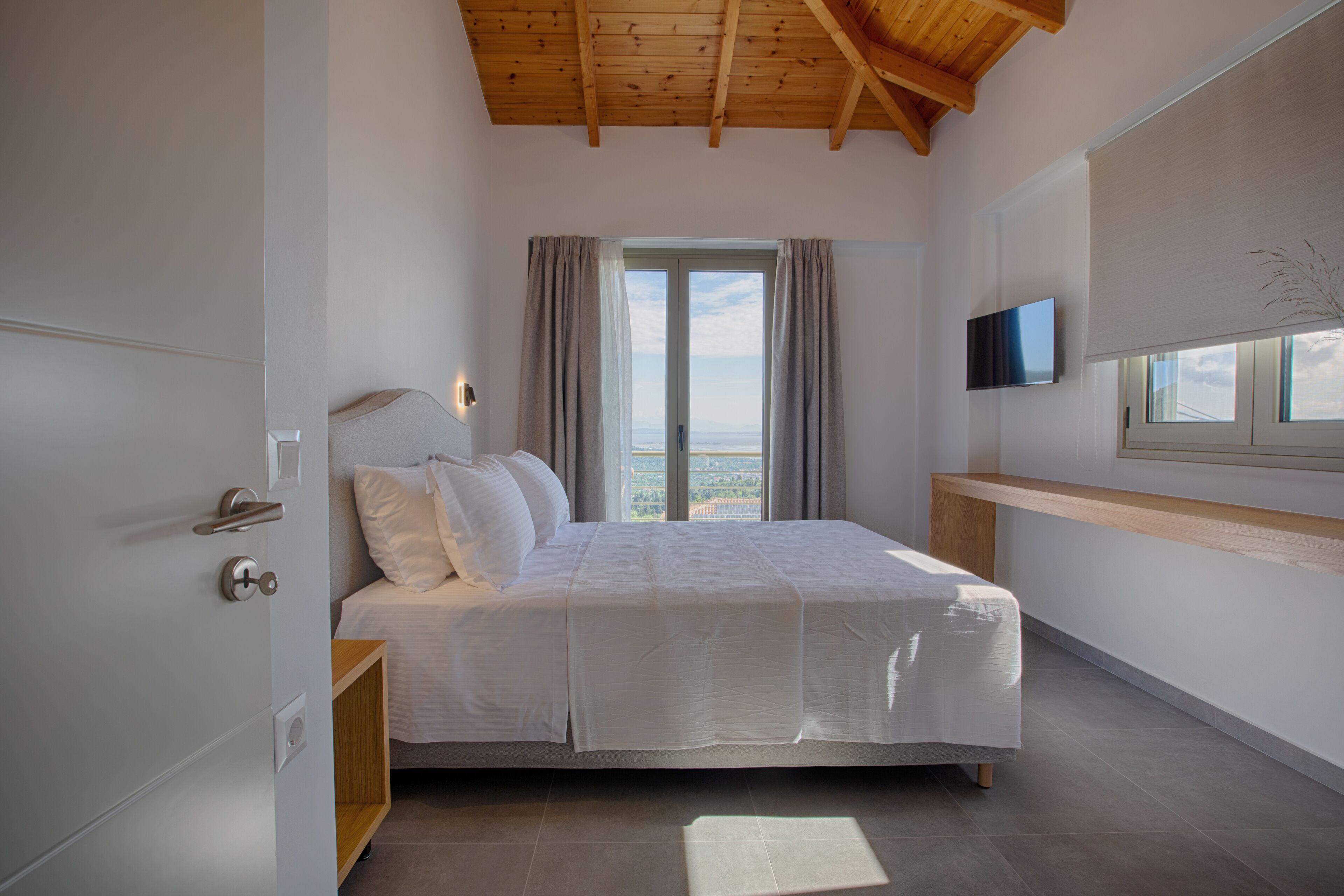 A bedroom with a bed, a TV mounted on the wall, a window with curtains, and a wooden ceiling.
