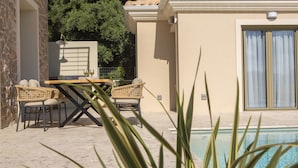 Un área junto a la piscina con una mesa y sillas, una piscina y un edificio con techo de tejas y un ventanal grande.