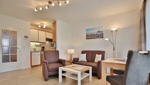 A modern living room with a sofa, armchair, coffee table, and dining table. There is a kitchen in the background with a microwave and cabinets.