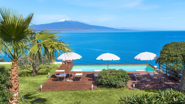 Zona de piscina con sillones y sombrillas, con vistas al mar azul y una montaña distante.