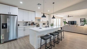 A modern kitchen with white cabinetry, a stainless steel refrigerator, a central island with bar stools, pendant lighting, and a large island sink. - Papago Haven, Private Oasis in Old Town (Scottsdale)