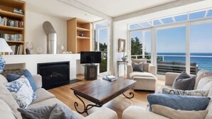 Een ruime woonkamer met een open haard, een televisie en een groot raam met uitzicht op zee.
