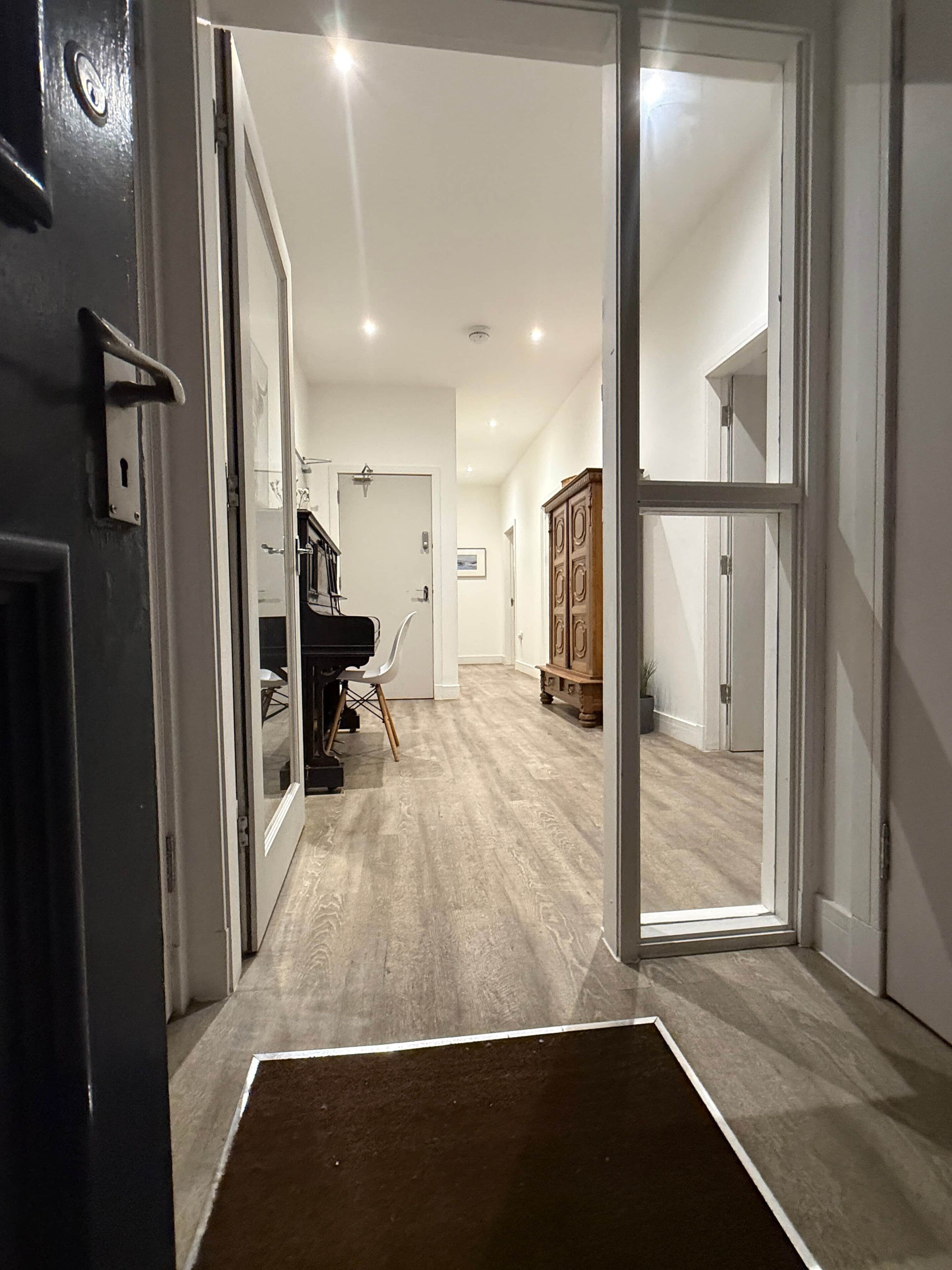 Un couloir menant à une pièce où se trouvent un piano, une armoire et une porte avec un interrupteur.