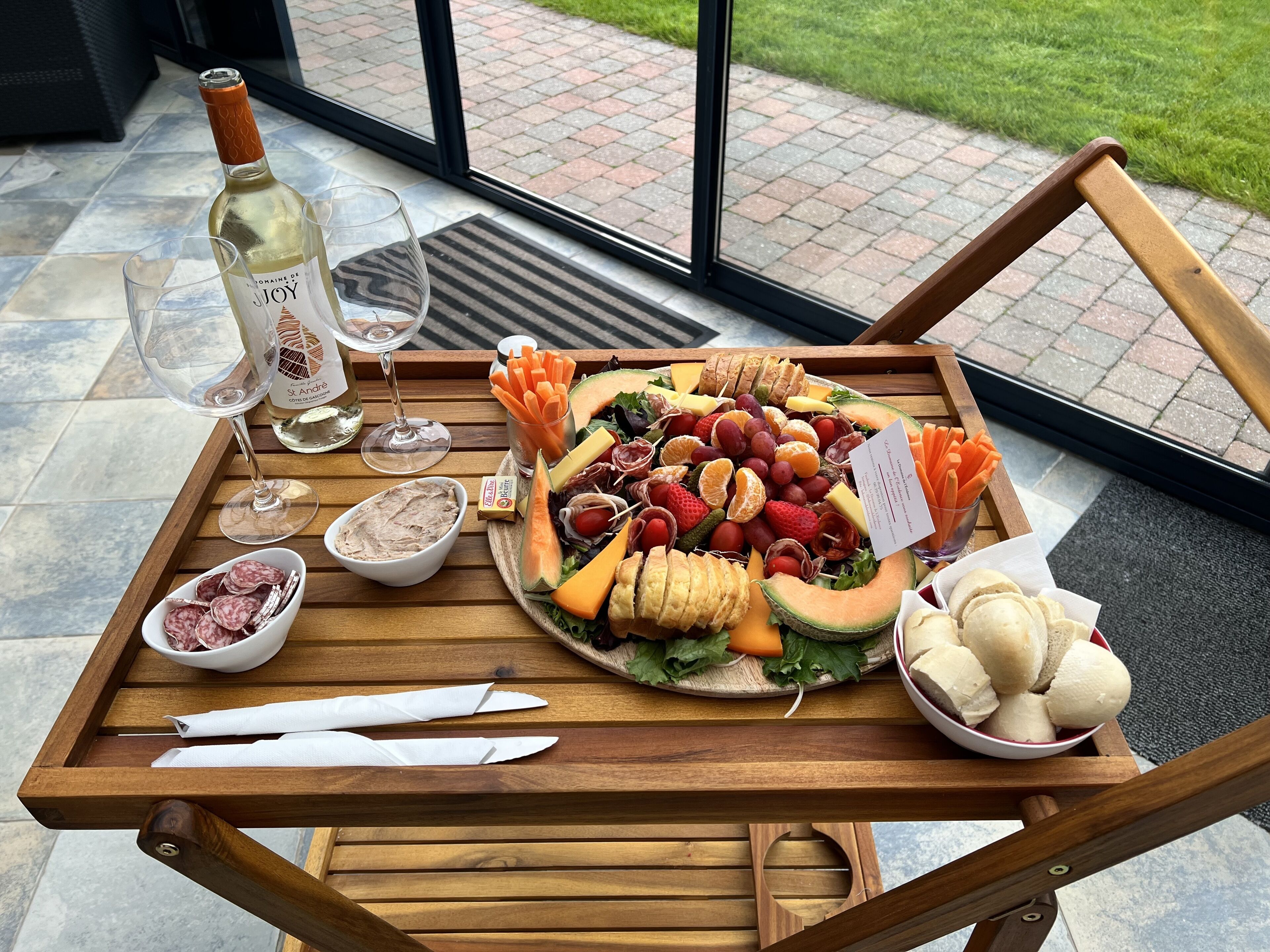 Un plateau en bois contenant une sélection de charcuteries, une bouteille de vin blanc et deux verres, le tout posé sur une terrasse dotée d’une porte vitrée.