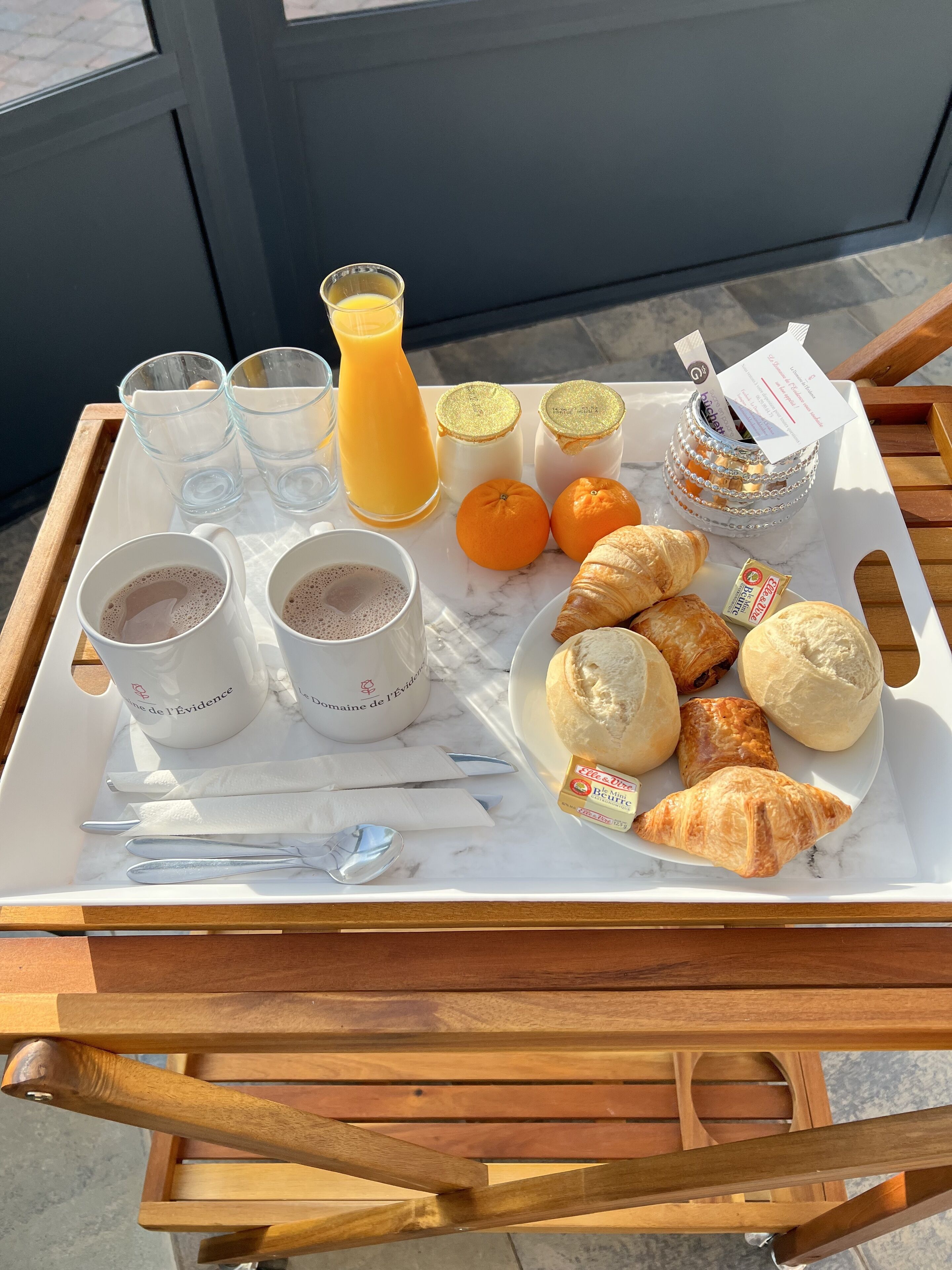 En bakke med croissanter, appelsinjuice, kaffe og et syltetøjsglas på et træbord.